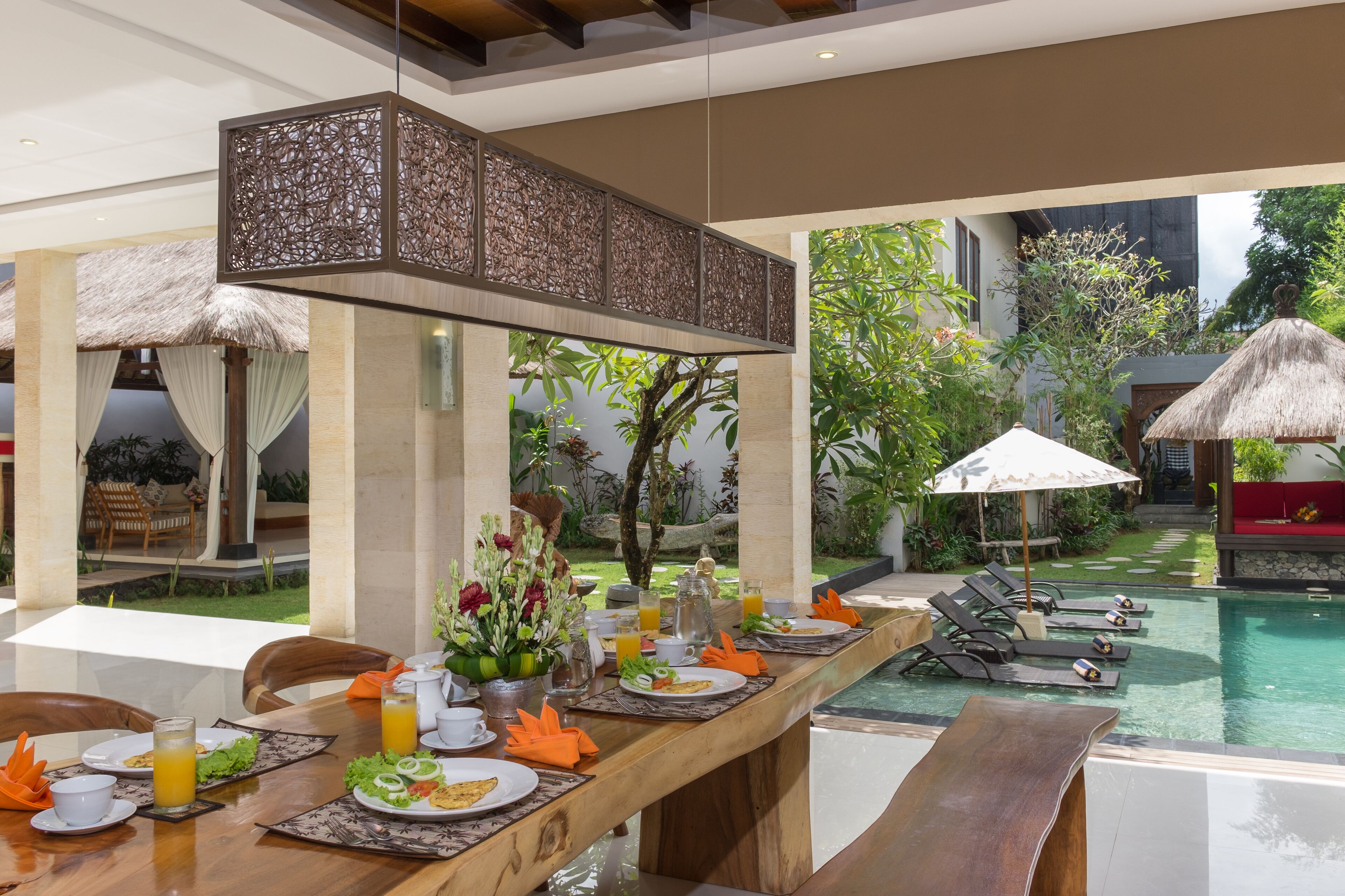 A dining area with a pool view, featuring a long wooden table set for breakfast, lounge chairs, and a thatched-roof structure.