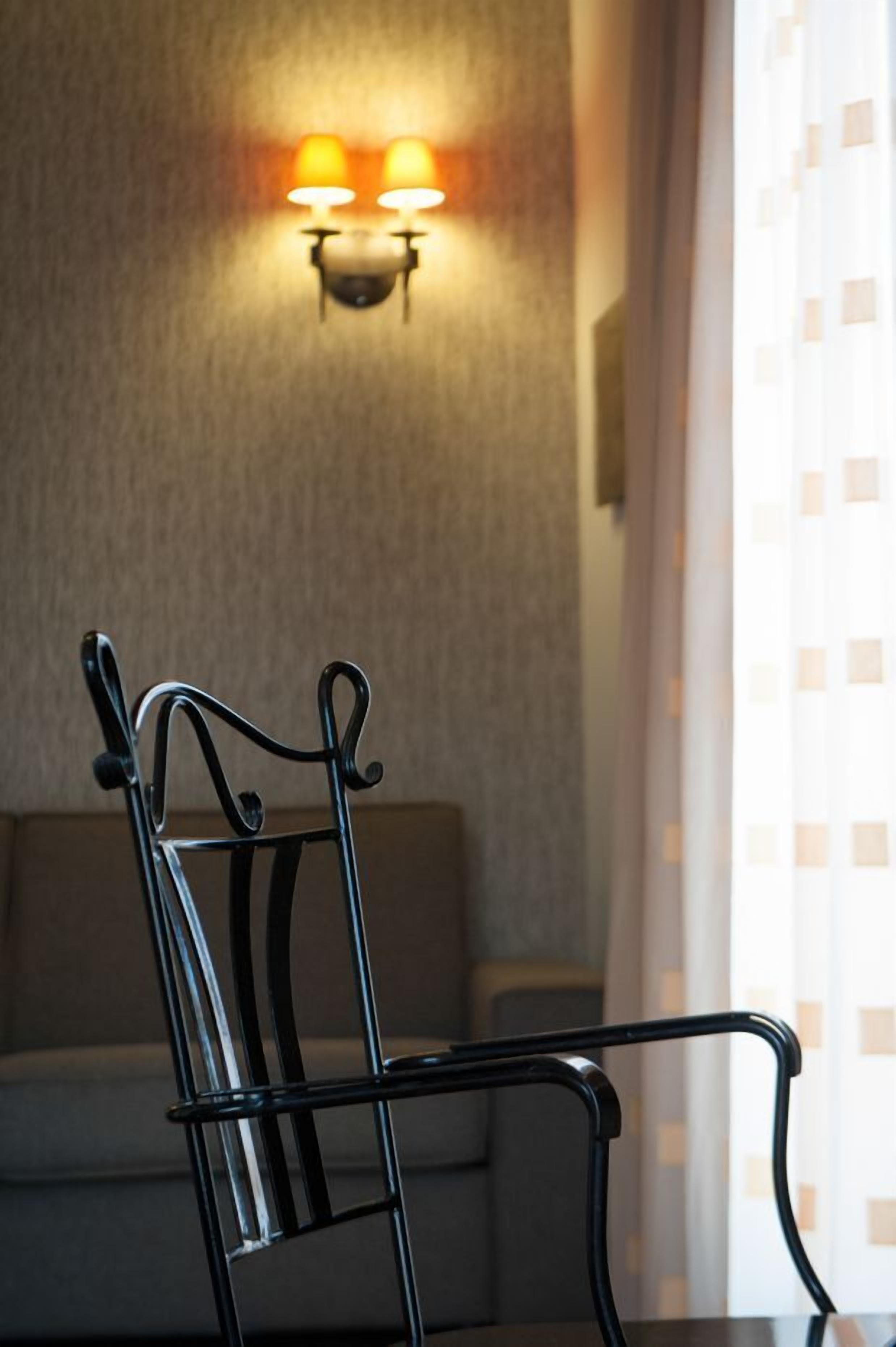 A dining area with a metal chair and a table, a wall-mounted light fixture, and a window with curtains.