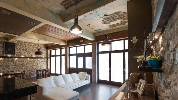 A spacious living area with a white sofa, a dining table, and a kitchen area with a stone wall. - Polys Guest House (Pydna-Kolindros)
