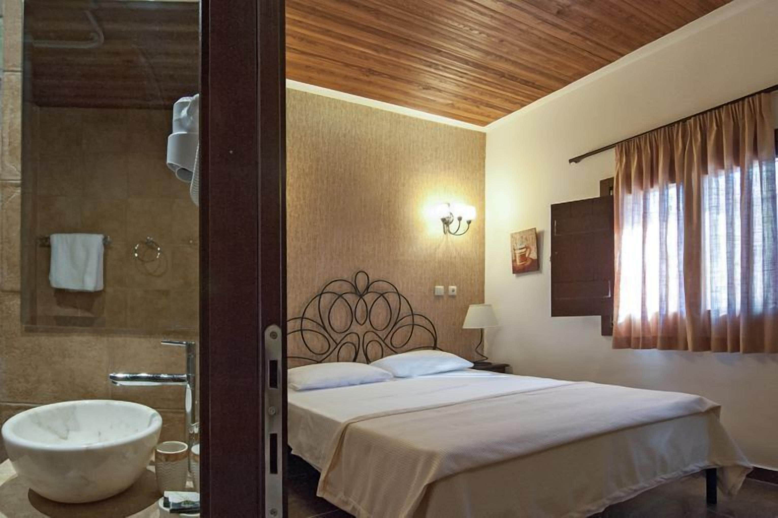 A hotel room with a wooden ceiling, a large bed with a decorative headboard, a bathroom with a round sink, and a window with curtains.