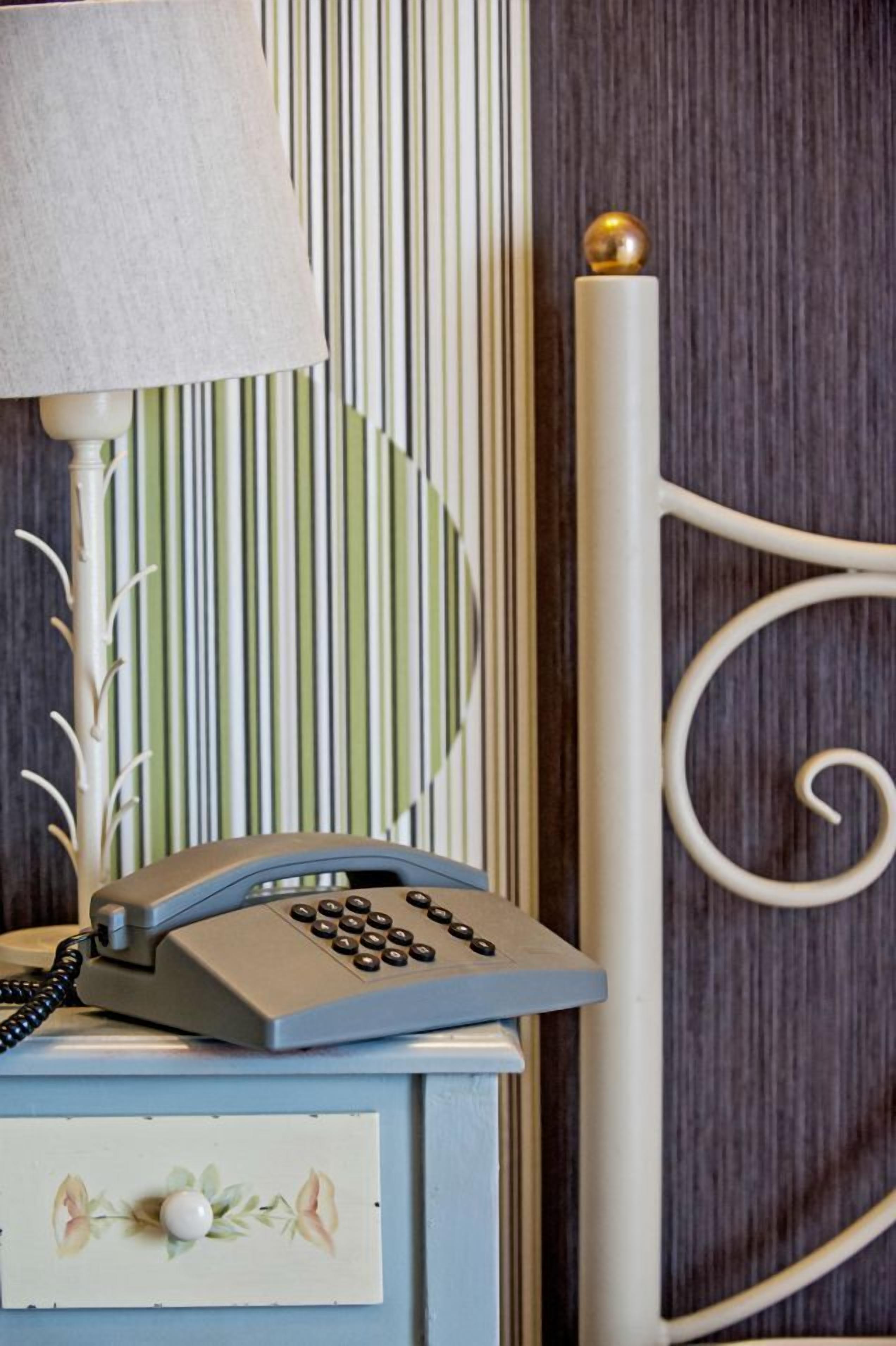 A vintage telephone on a bedside table with a lamp, a decorative wall panel, and a metal bed frame.