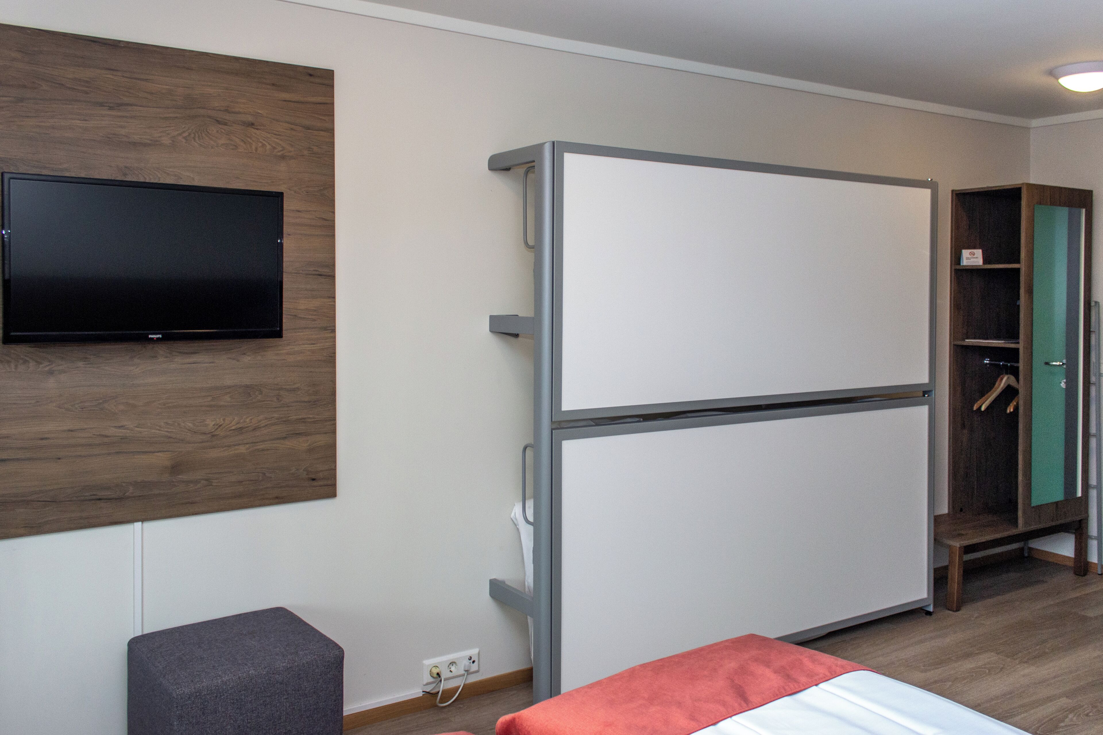 Habitación de hotel compacta con televisión de pantalla plana montada en un panel de madera, minibar blanco, otomán gris pequeño y una cama con un detalle en rojo.