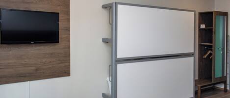 Habitación de hotel compacta con televisión de pantalla plana montada en un panel de madera, minibar blanco, otomán gris pequeño y una cama con un detalle en rojo.