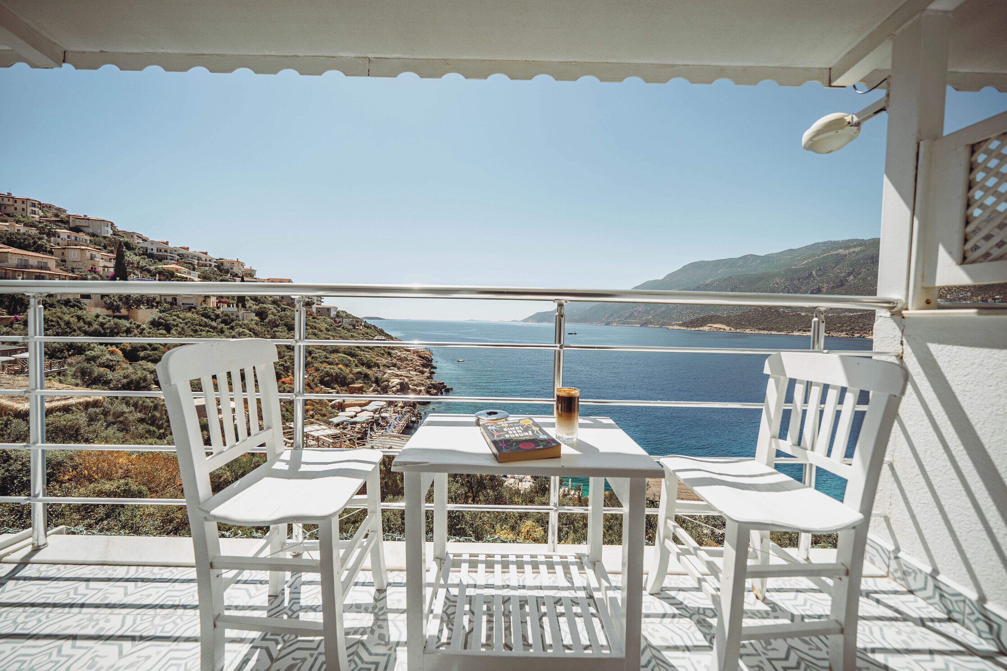 A balcony with white chairs and a table, overlooking a coastal landscape with buildings and the sea.