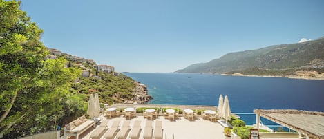 A poolside area with lounge chairs and umbrellas, overlooking a body of water and a mountainous landscape.