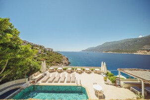 Zona junto a la piscina con sillas de descanso y sombrillas, con vista a una masa de agua y un paisaje montañoso.