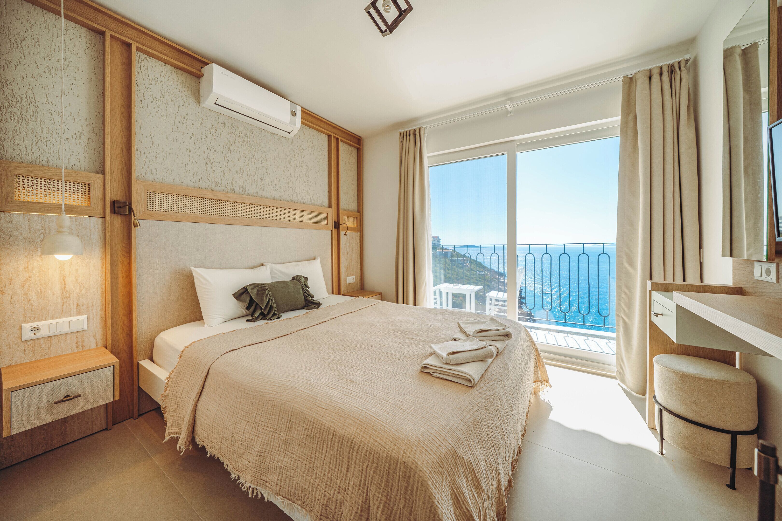 A hotel room with a bed, bedside table, a desk with a chair, a large window with a view, and an air conditioning unit.