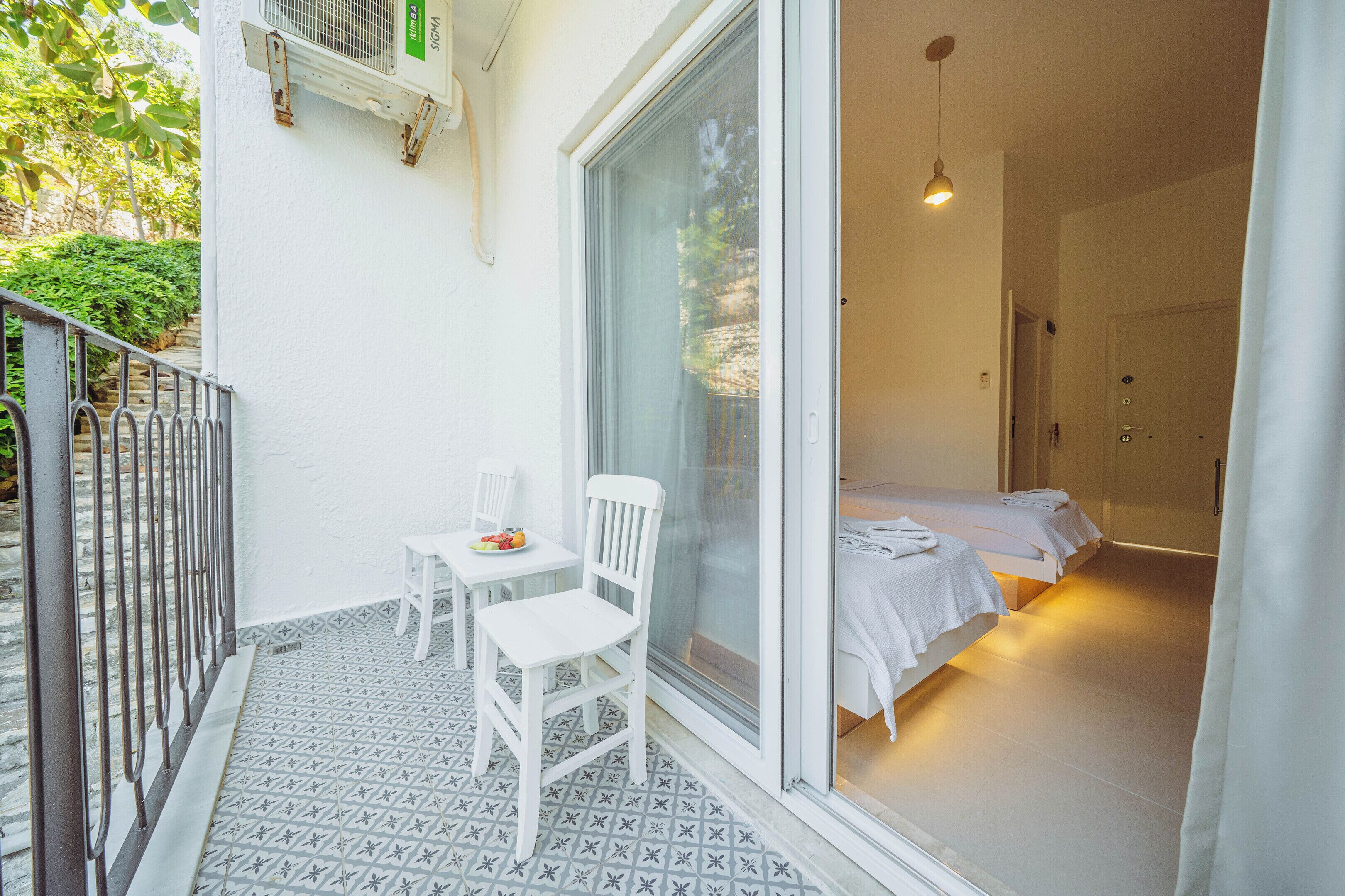 Un balcón con una silla y una mesa blancos, una puerta corrediza de vidrio que da a una habitación con dos camas y una lámpara colgante.