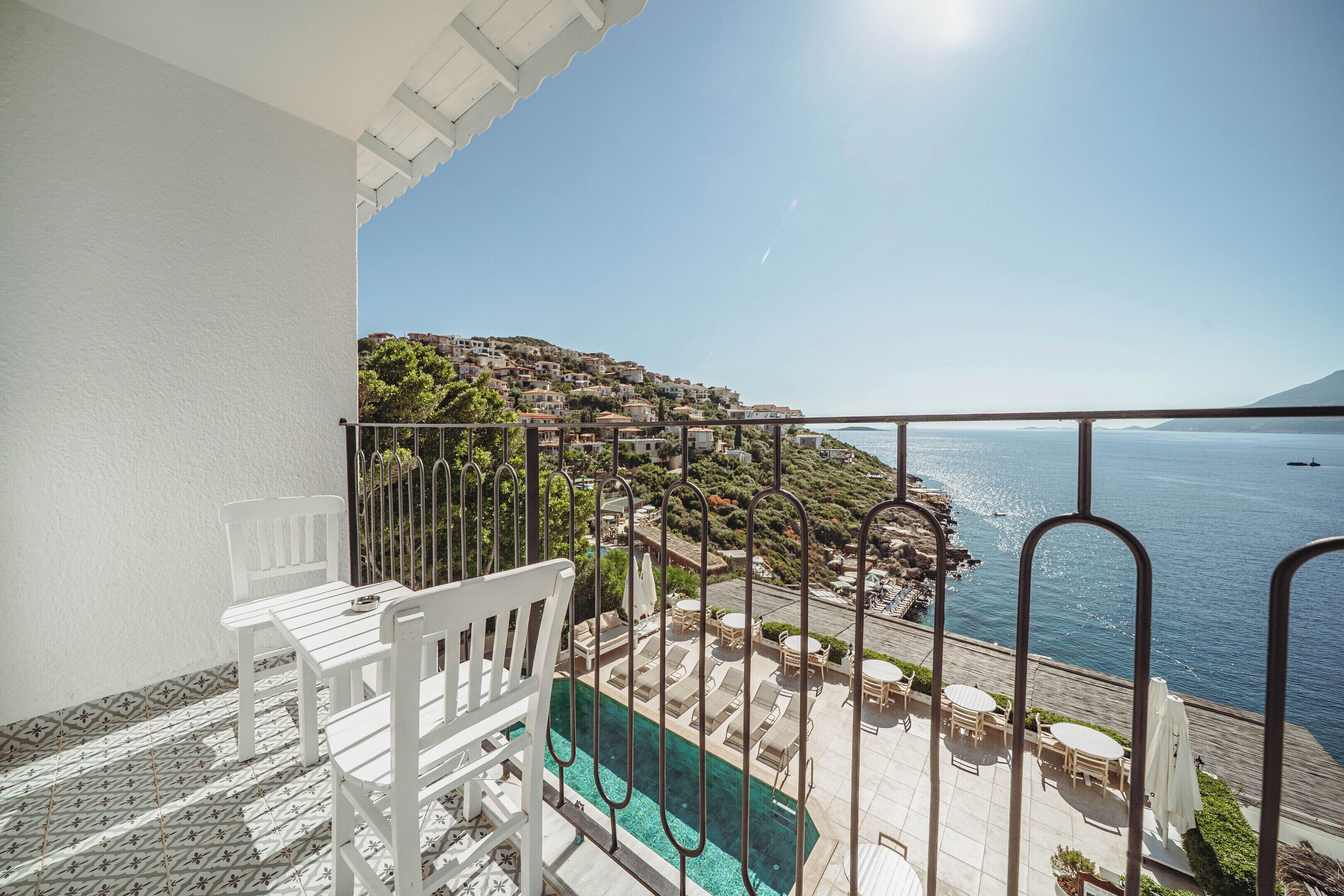 A balcony with a pool, white chairs, and a view of the sea and hillside buildings.