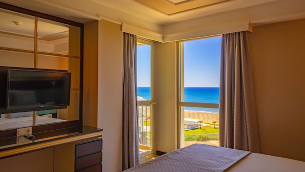 Une chambre d’hôtel équipée d’un lit, d’une télévision, d’un bureau et d’une grande fenêtre donnant sur la plage et l’océan.