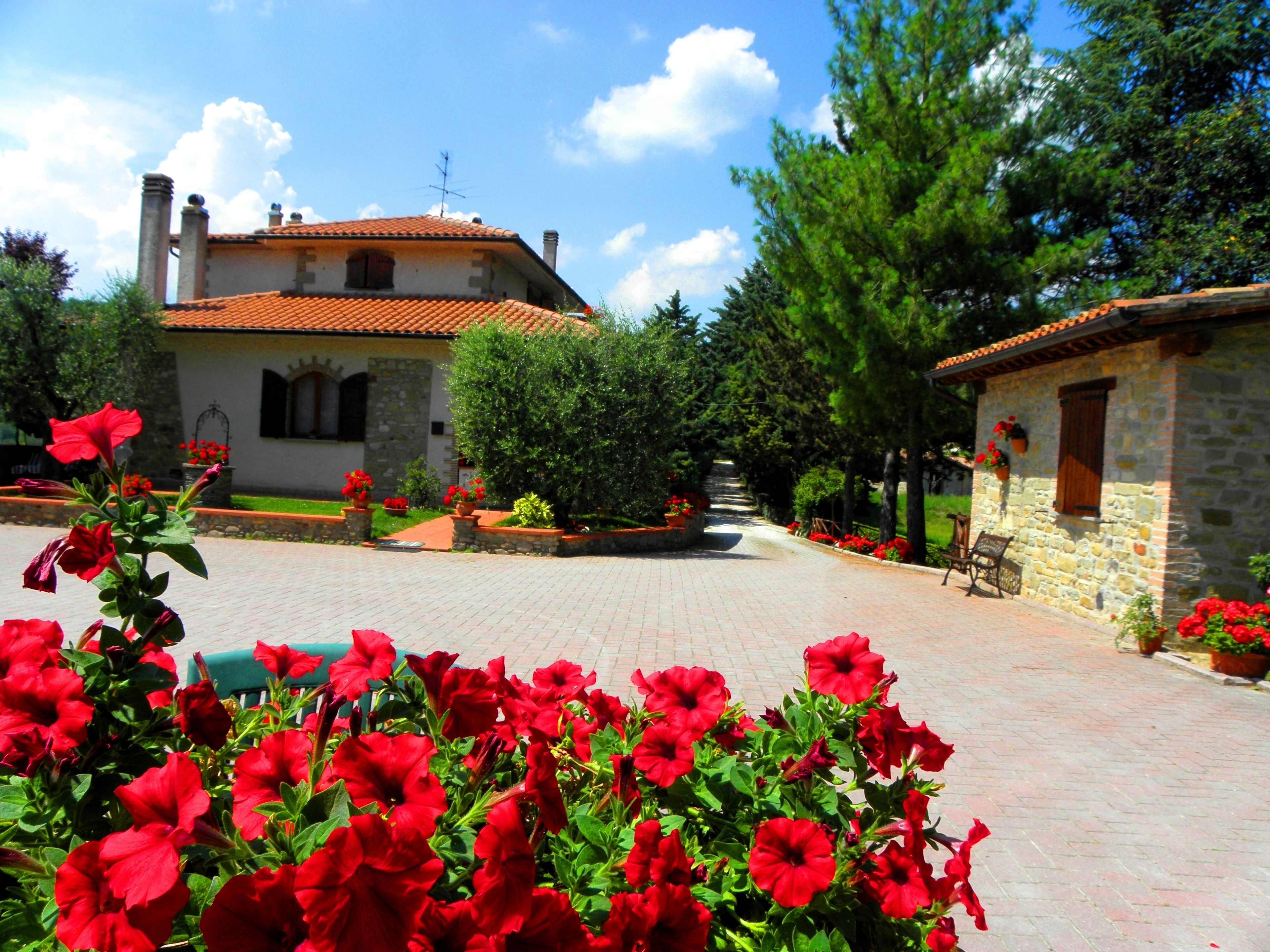 A stone building with a tiled roof, a paved courtyard, and red flowers in the foreground.