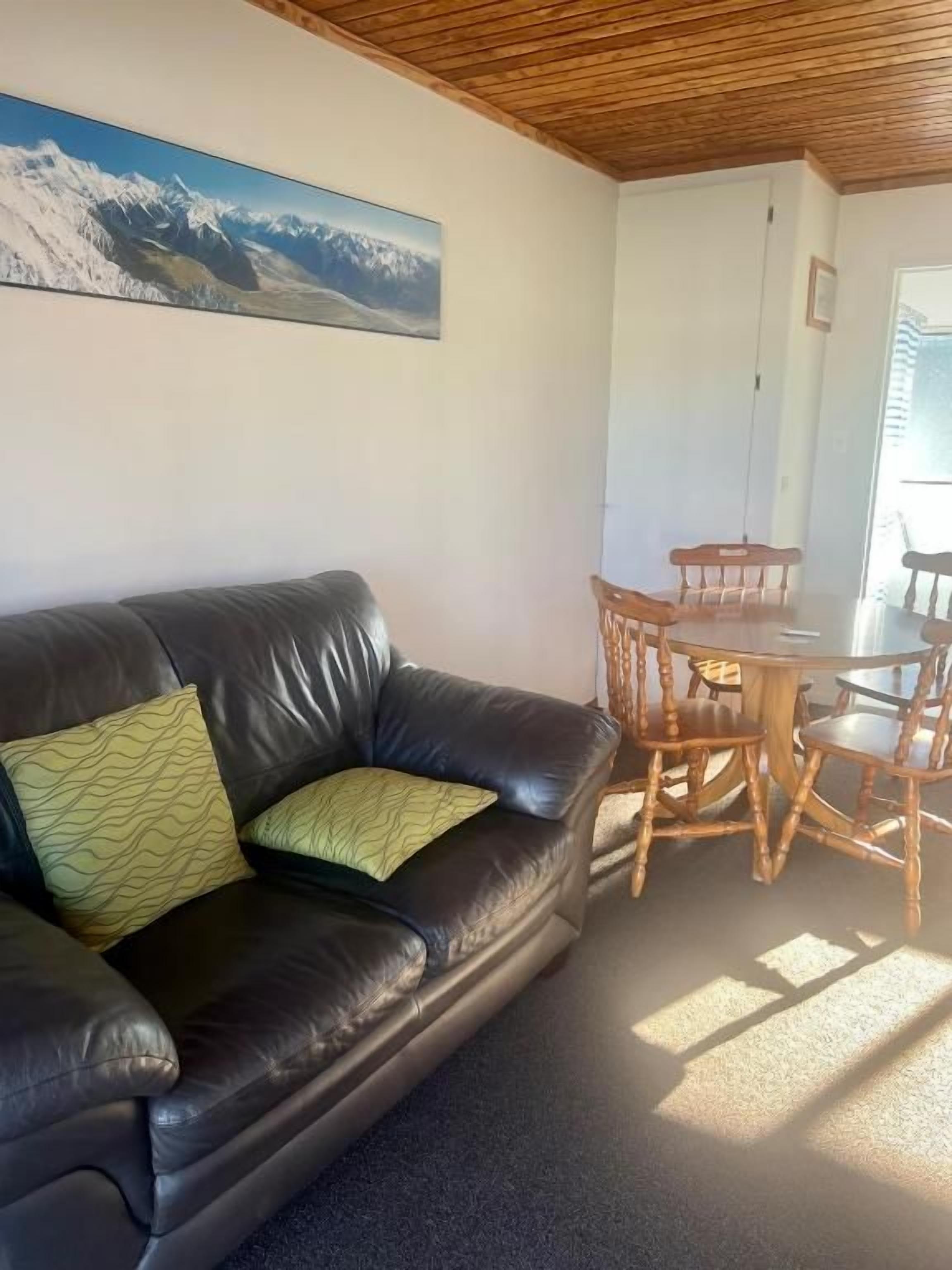 Una sala de estar con un sofá de cuero, una mesa de comedor de madera con sillas y un cuadro de un paisaje montañoso en la pared.