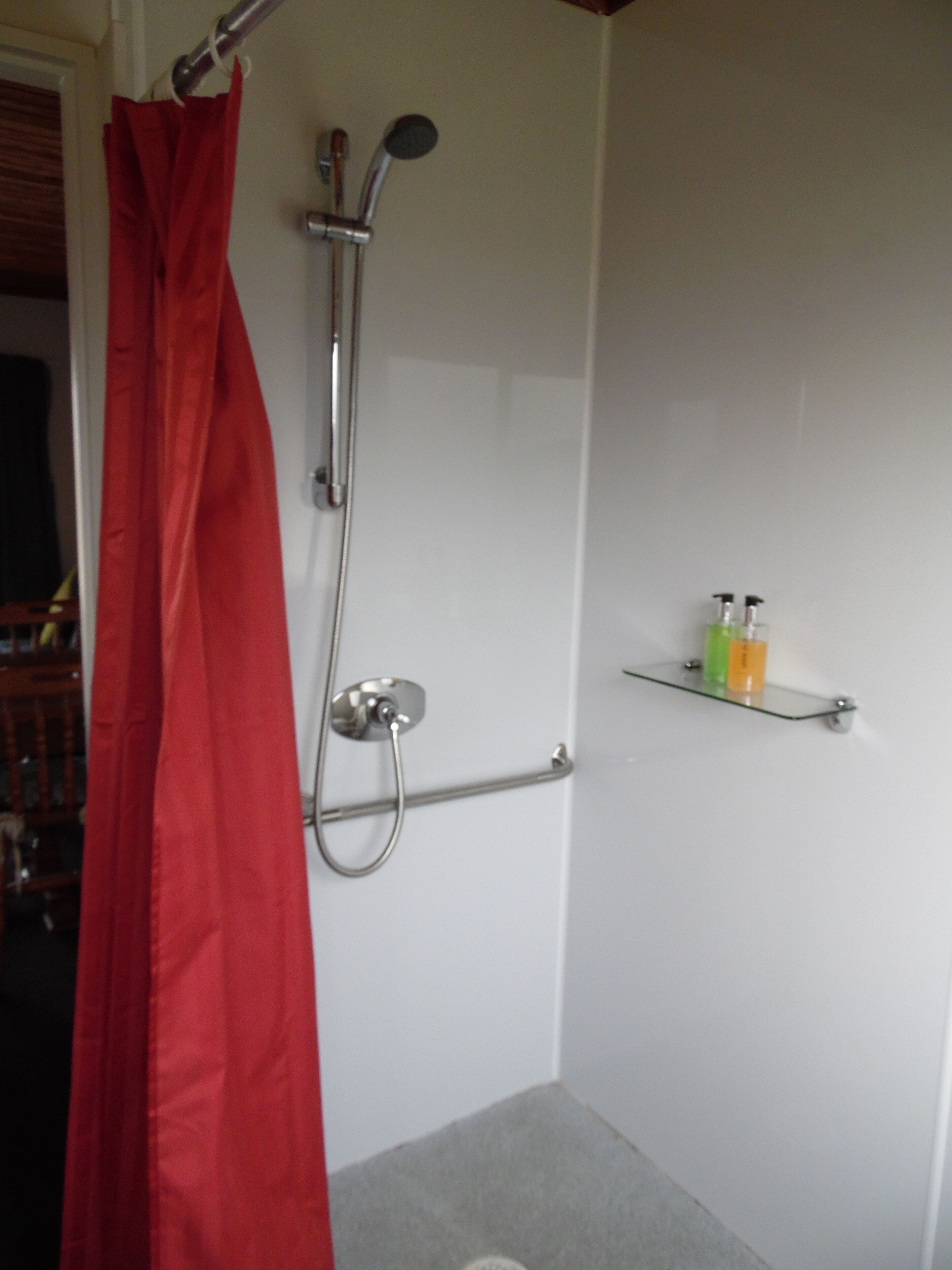 A shower with a red curtain, a glass shelf with toiletries, and a towel rack.