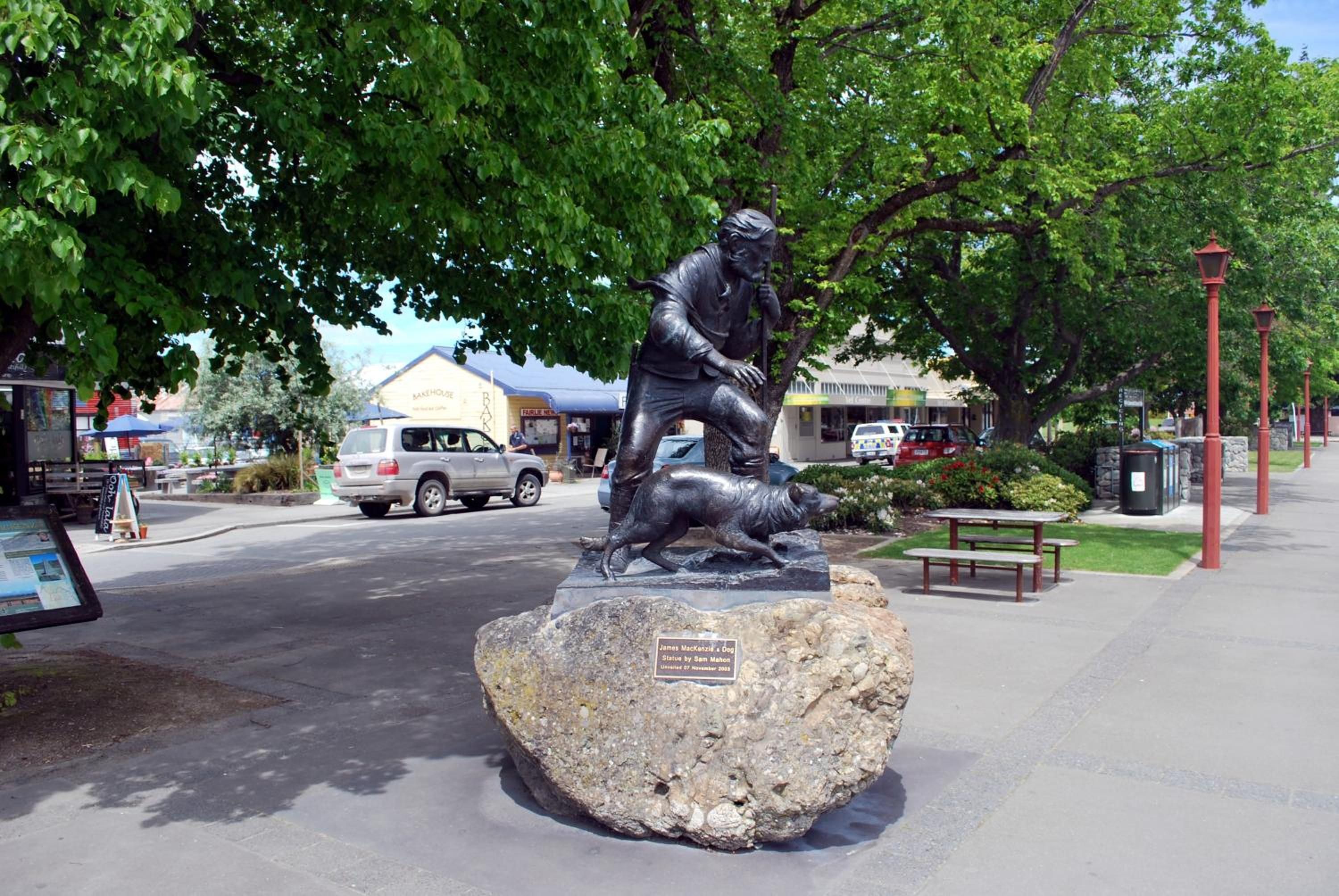 Una estatua de un hombre con un perro sobre una roca, un banco y una calle con coches y edificios.