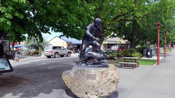 Una estatua de un hombre con un perro sobre una roca, un banco y una calle con coches y edificios.