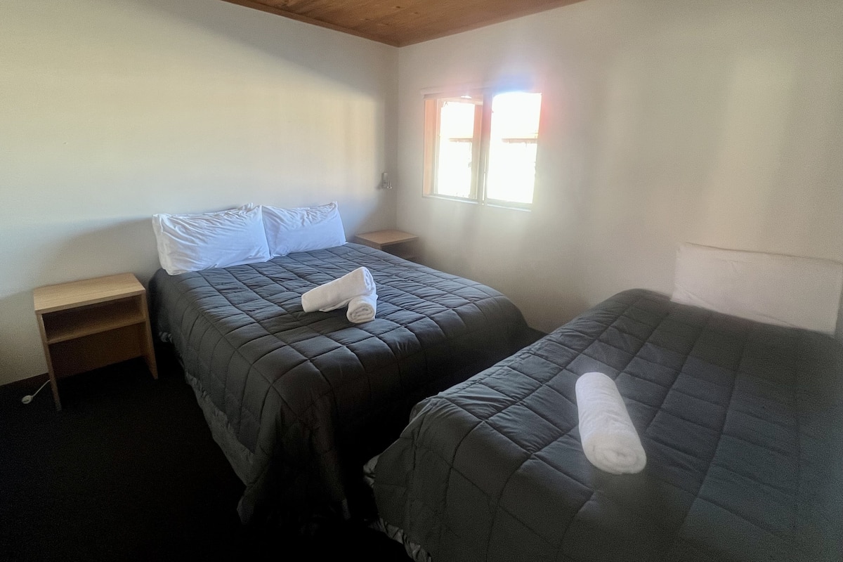 A room with two beds, each with a dark quilted bedspread and white pillows. There is a small wooden nightstand on the left side of the beds, and a window on the right wall allowing natural light to enter.