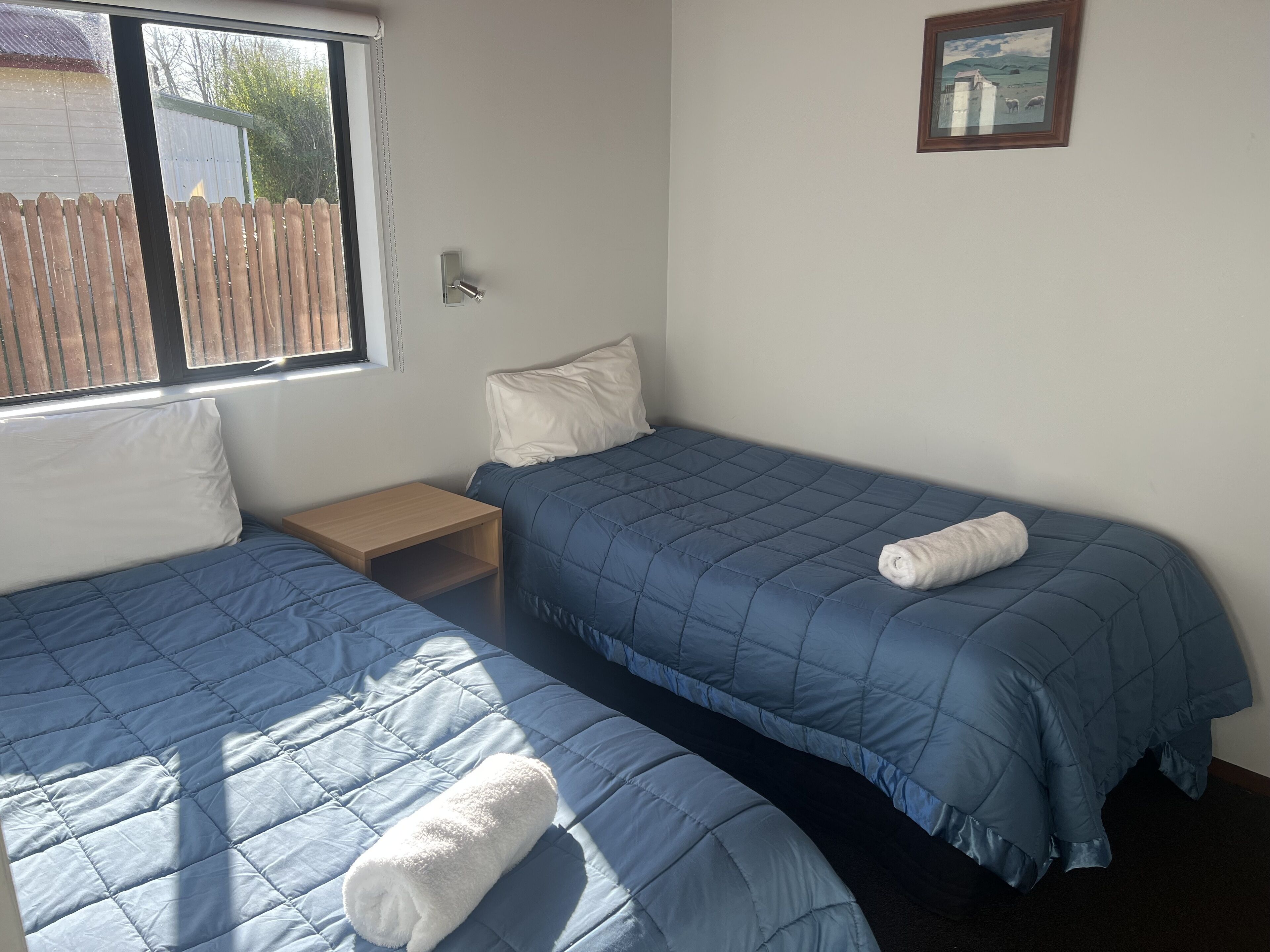 Une chambre avec deux lits, chacun recouvert d’une courtepointe bleue matelassée et agrémenté d’un oreiller blanc. Une table de chevet en bois se trouve entre les deux lits, et un tableau encadré est accroché au mur.