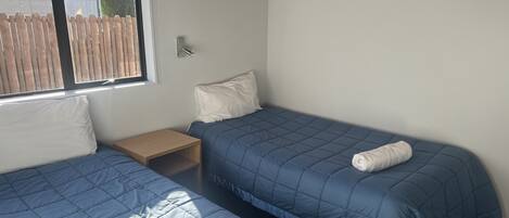 Une chambre avec deux lits, chacun recouvert d’une courtepointe bleue matelassée et agrémenté d’un oreiller blanc. Une table de chevet en bois se trouve entre les deux lits, et un tableau encadré est accroché au mur.
