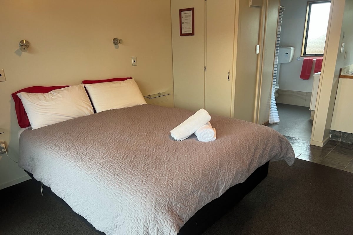 A neatly made bed with a beige quilt and two pillows, a towel on the bed, and a visible bathroom with a toilet and towel rack.