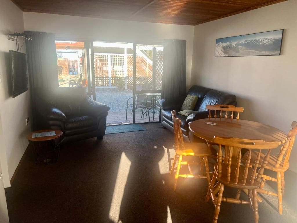 Une pièce comprenant un canapé, une table à manger avec des chaises, un téléviseur fixé au mur et une vue sur l’extérieur, visible à travers la porte.