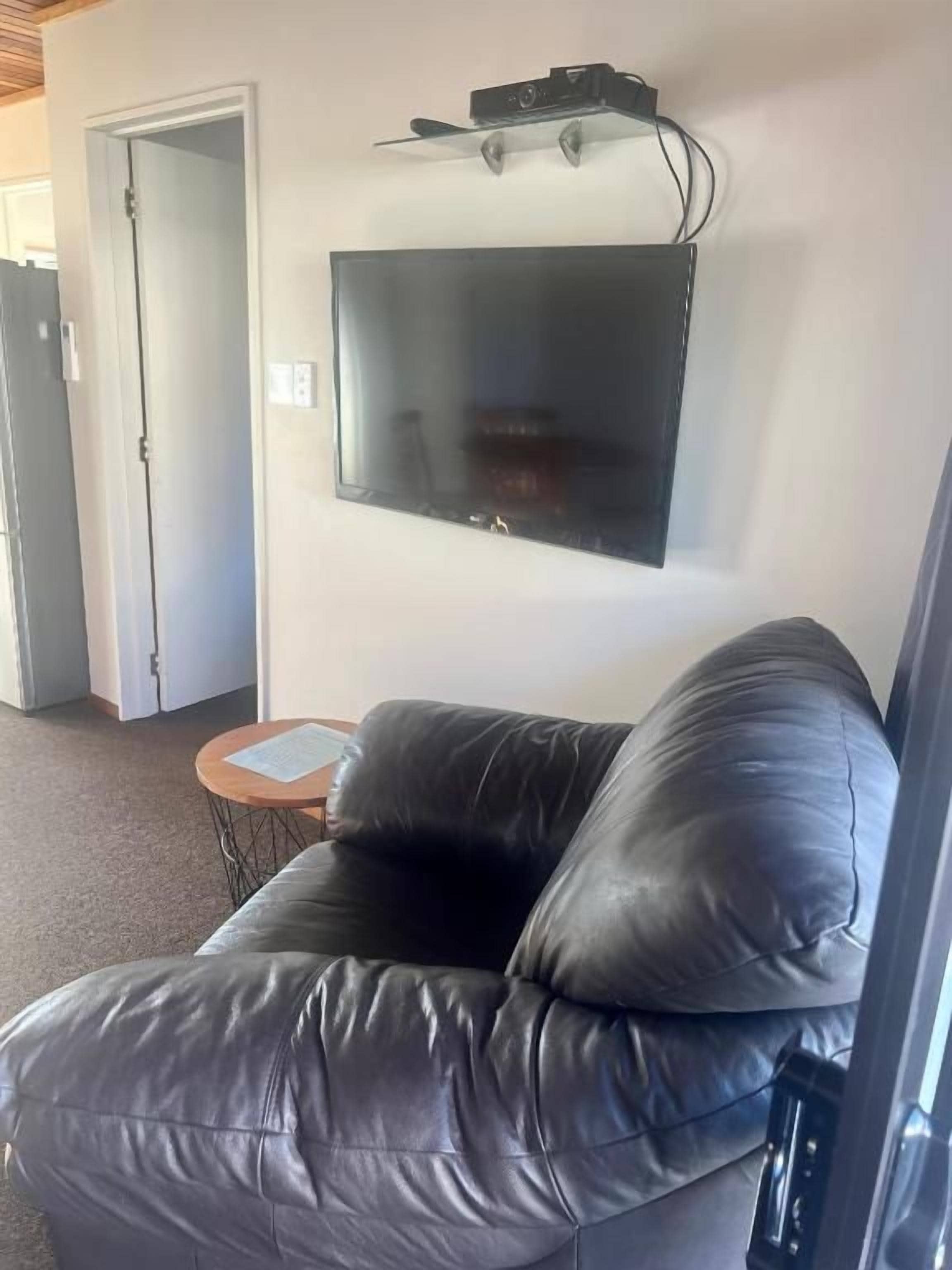 Un salon avec un canapé en cuir noir, un téléviseur à écran plat fixé au mur et une petite table ronde sur laquelle se trouve un document.