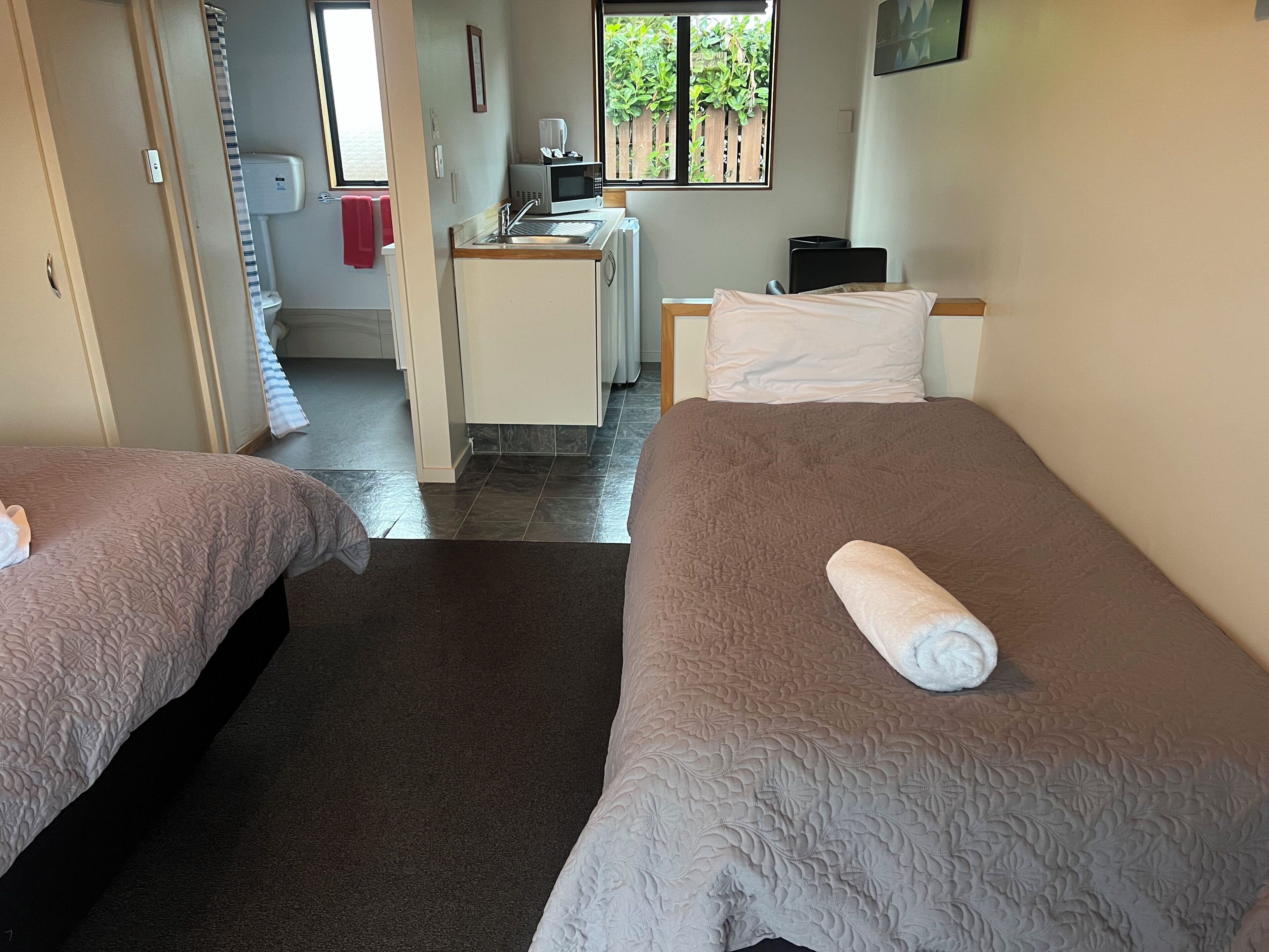 A hotel room with two beds, a desk, a window with a view of greenery, and a bathroom visible through an open door.