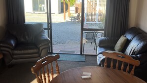 Ett rum med ett träbord och stolar, en soffa i läder och en utsikt genom en glasdörr till ett utomhusområde med en uteplats och ett staket.