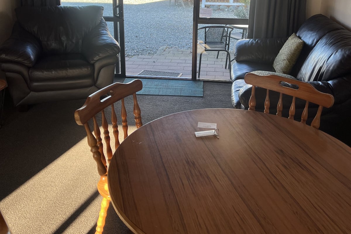 A room with a wooden table and chairs, a leather sofa, and a view through a glass door to an outdoor area with a patio and a fence.
