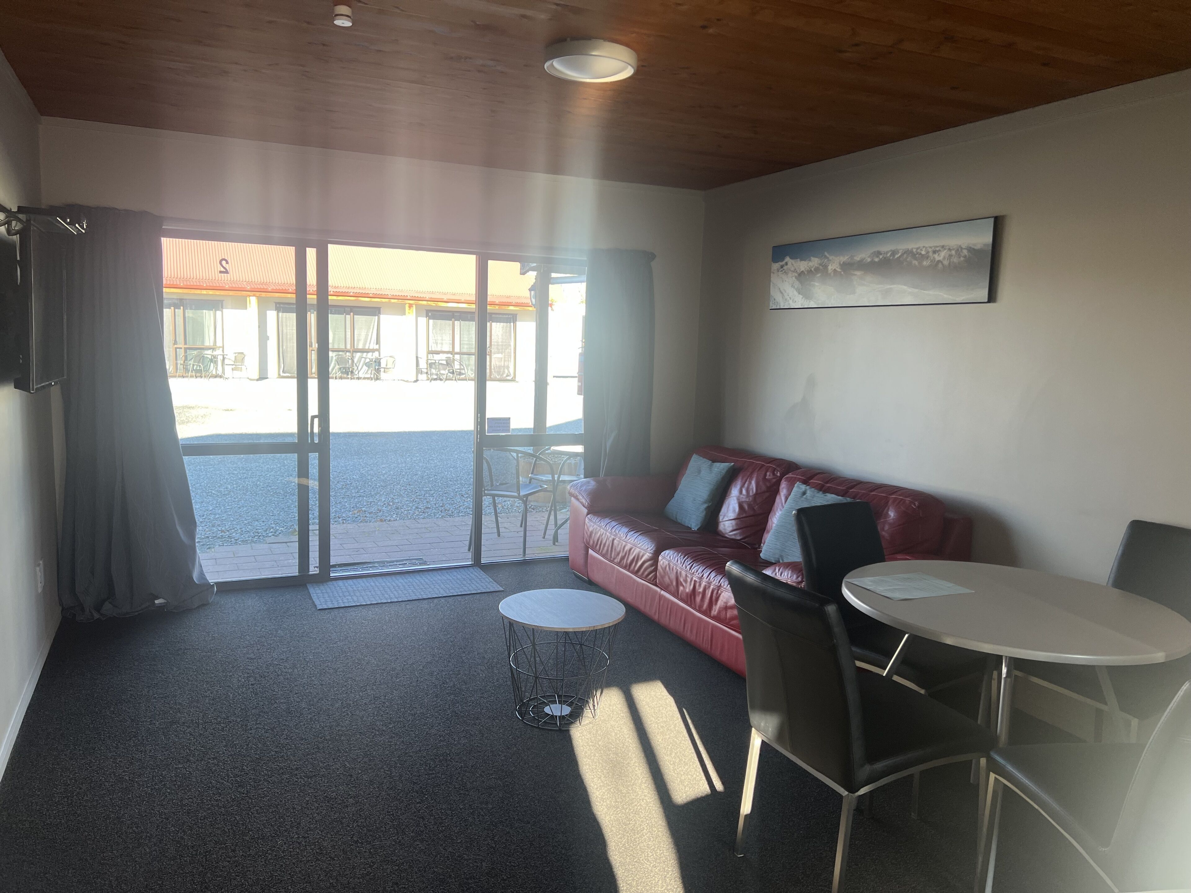 A room with a red leather sofa, a small round table, and a chair. There is a sliding glass door leading to an outdoor area with tables and chairs.