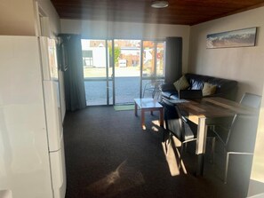 A room with a sofa, a table, and a refrigerator. There is a window with curtains, and a picture on the wall.