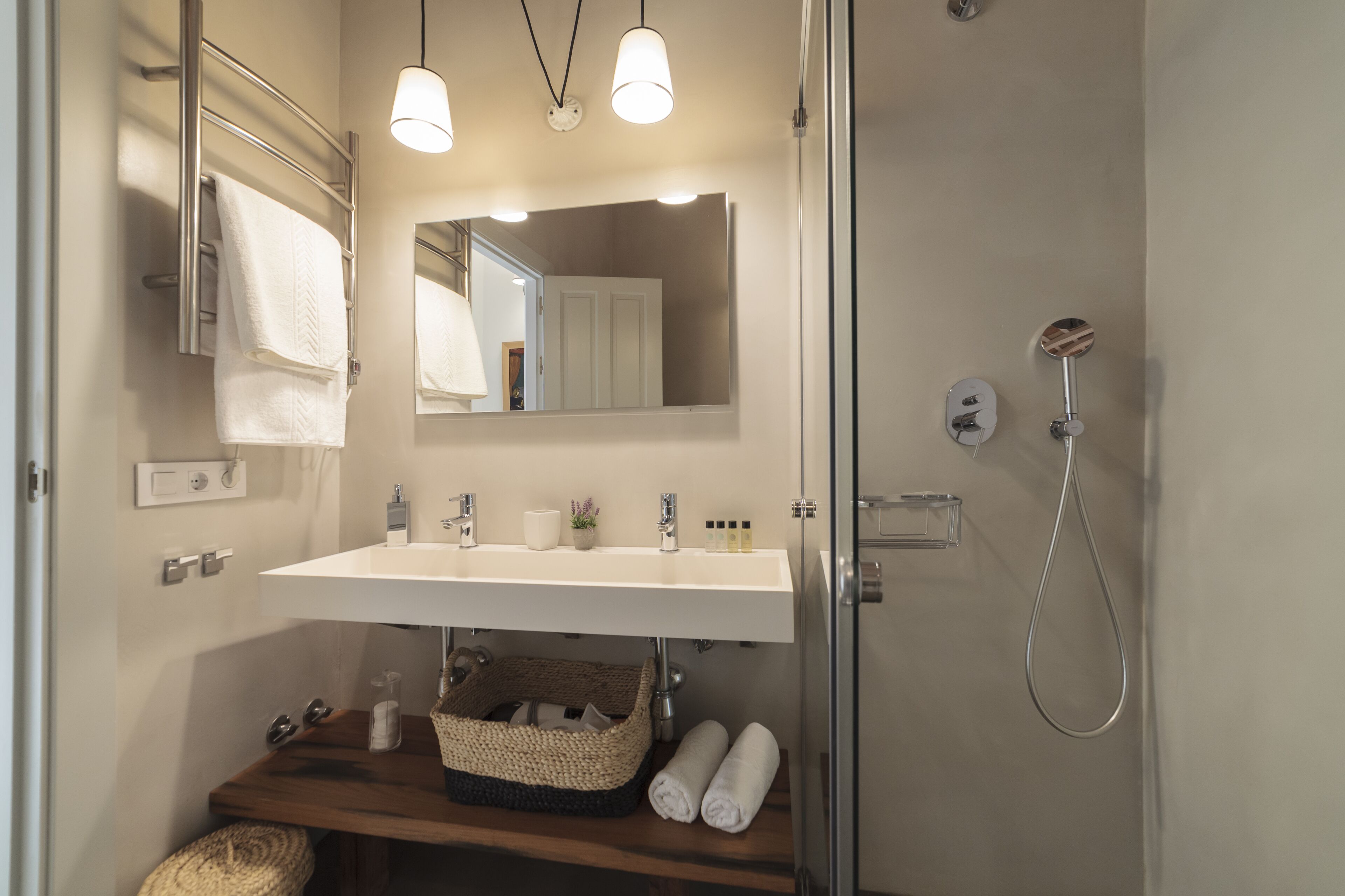 A bathroom with a double sink, a mirror, a towel rack, a shower, and a wooden shelf with towels and toiletries.