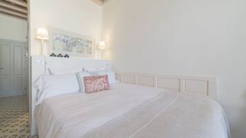 Una cama bien hecha con cabecera blanca y una sola almohada decorativa. Encima de la cama hay una obra de arte enmarcada, y a la izquierda se ve una mesita con una lámpara.