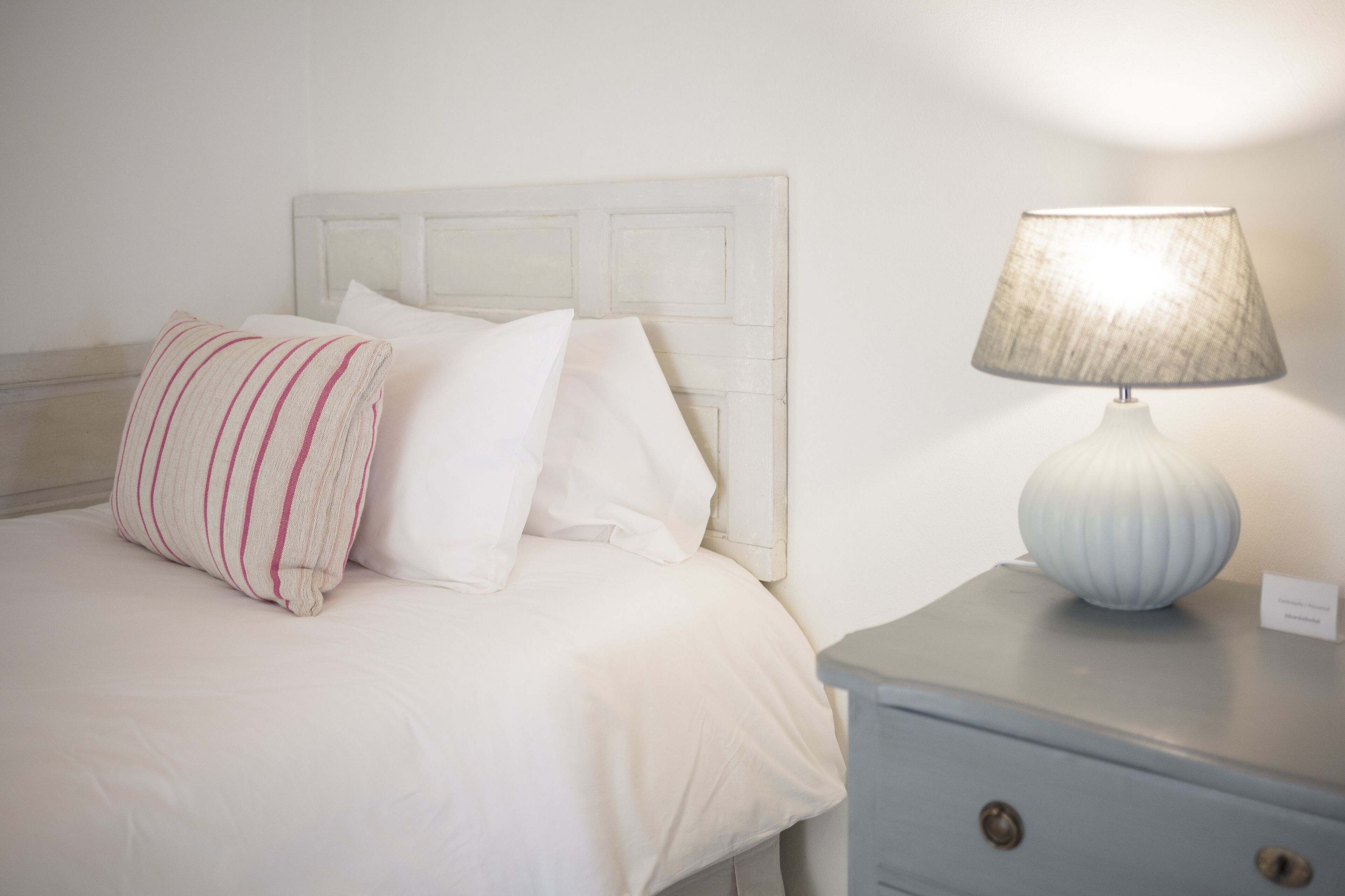 Una cama bien hecha con ropa blanca y almohadas a rayas, una lámpara de noche y un mueble auxiliar con cajones.