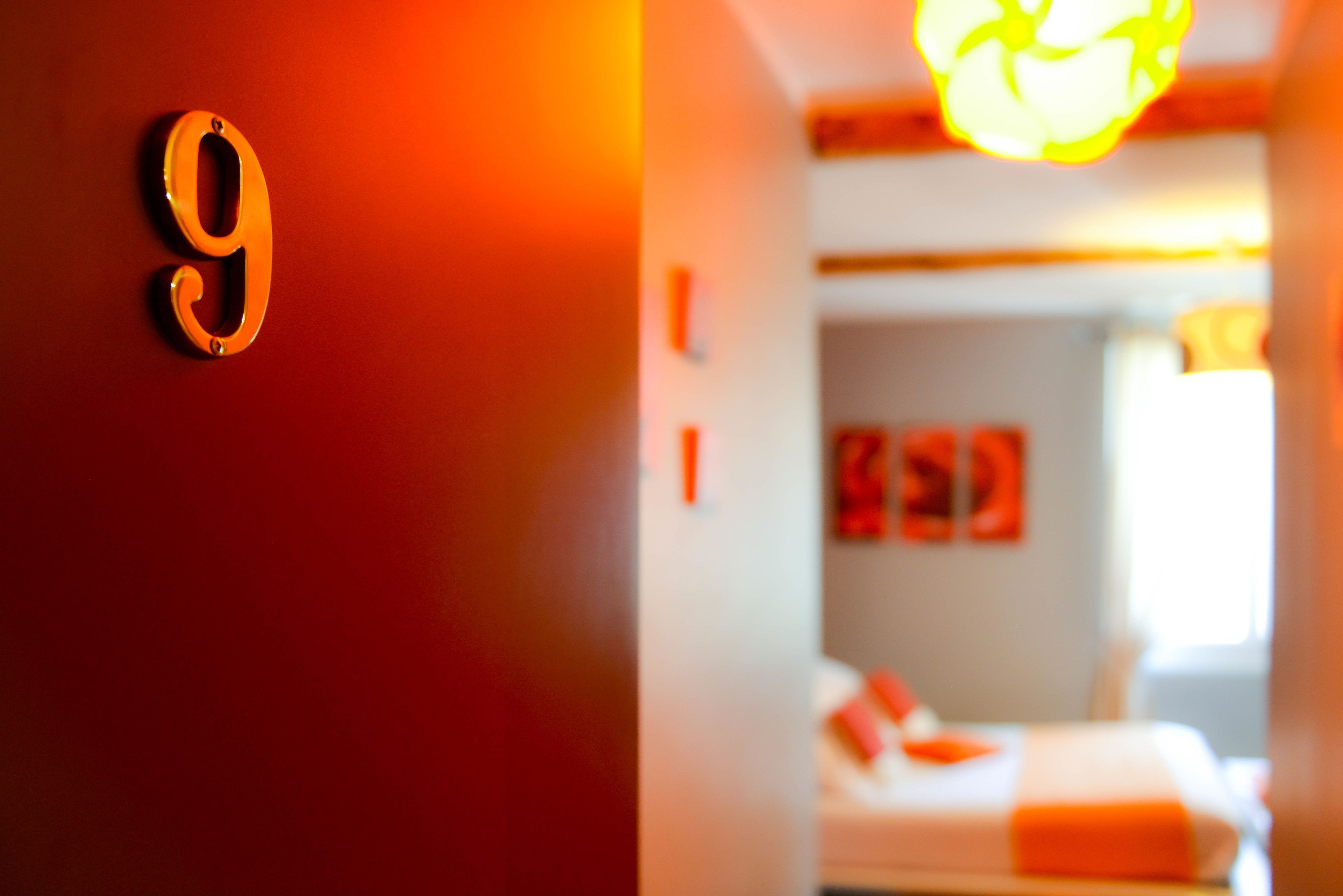A hotel room with a large '9' sign on the wall, a bed with orange bedding, and a bright light fixture above the bed.