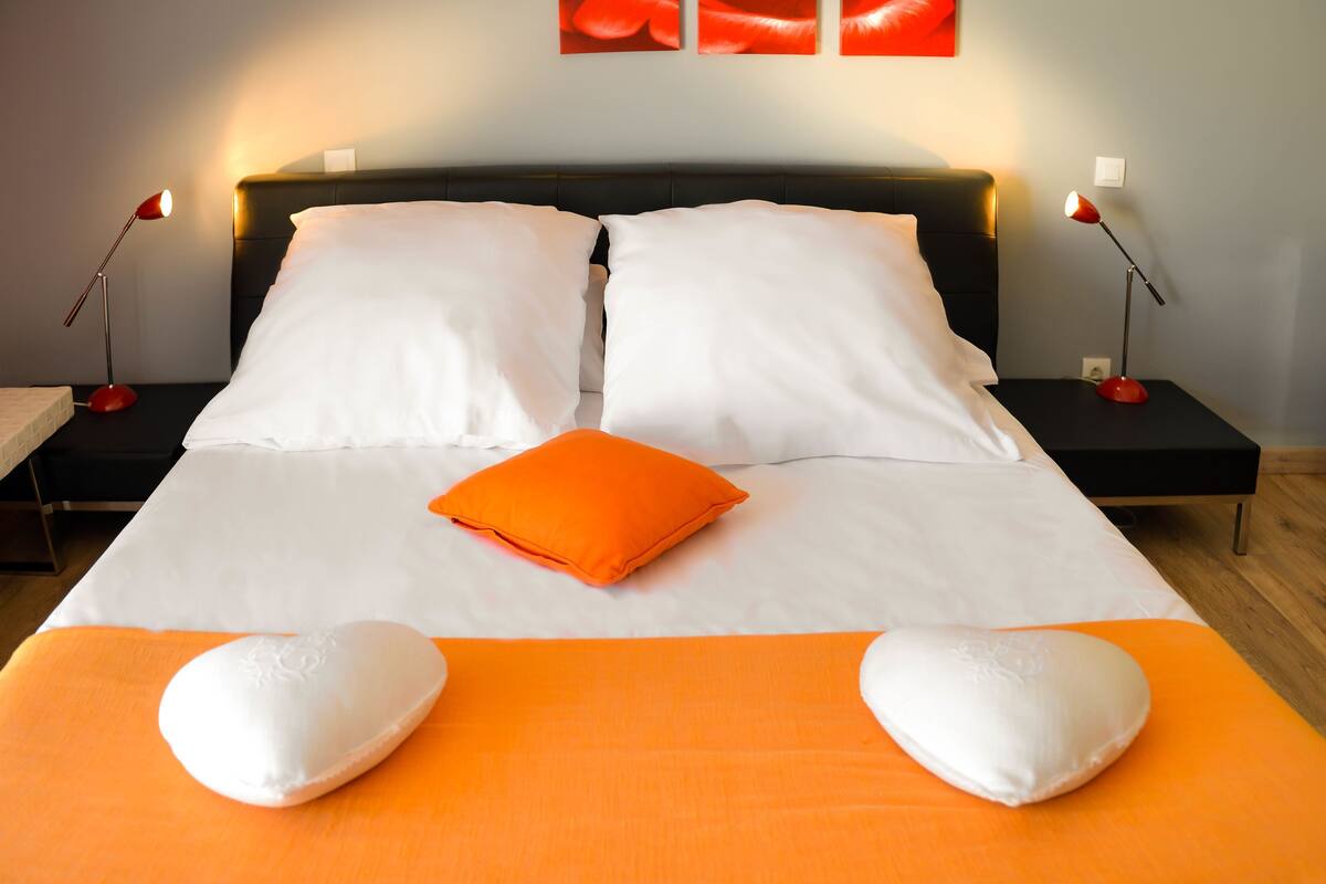Un lit bien fait, avec une literie blanche et orange, deux tables de chevet avec des lampes, et un mur orné d’un tableau abstrait rouge.