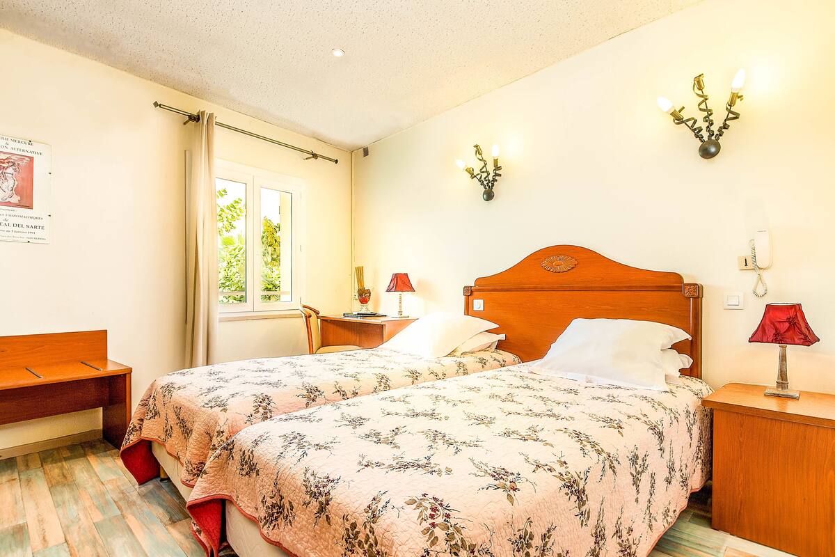 Un lit double avec une literie à motifs floraux, une table de chevet en bois avec une lampe, un banc en bois et une fenêtre avec des rideaux.