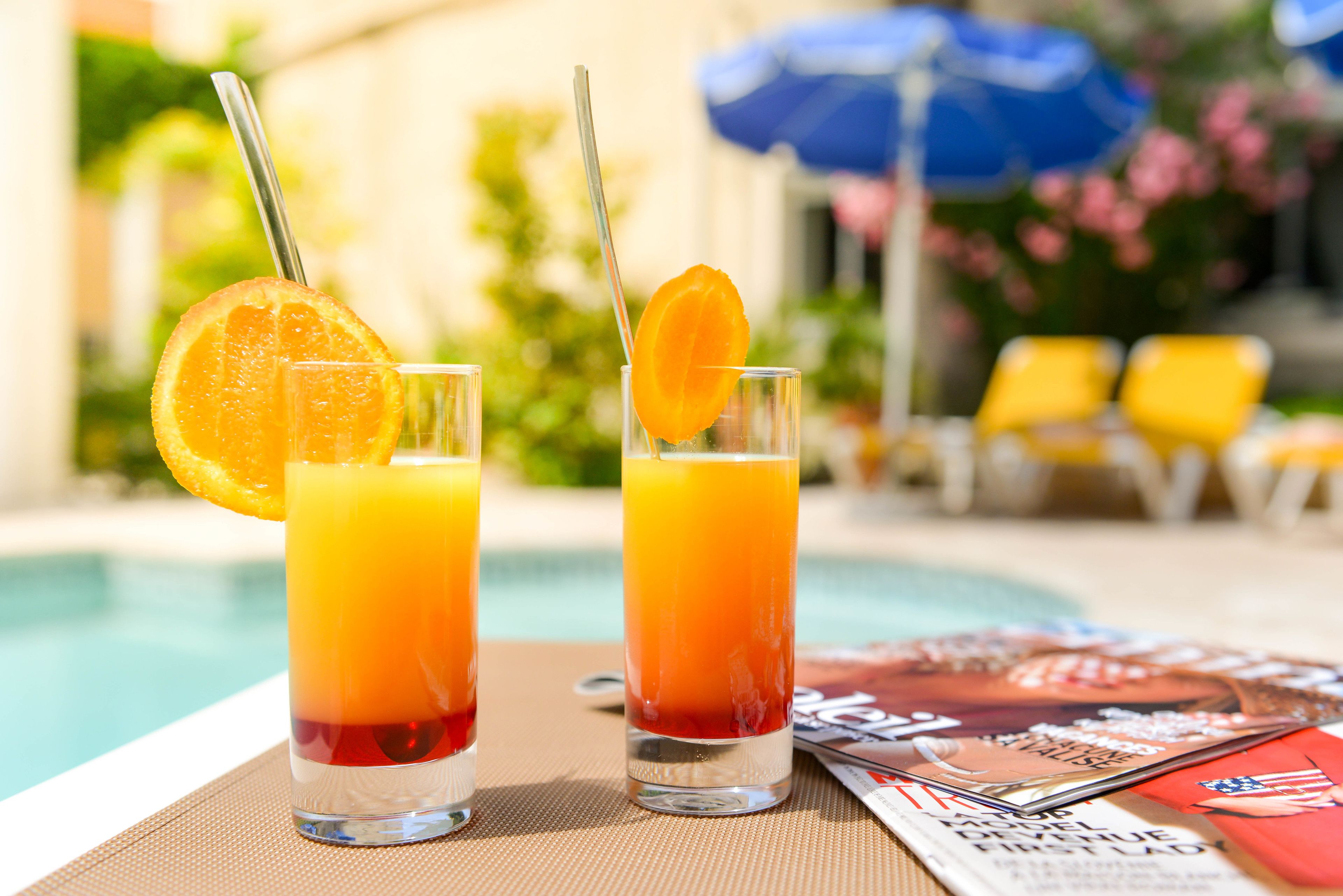 Two glasses of orange juice with orange slices on the rim, placed on a table near a poolside area with yellow chairs and a blue umbrella in the background.