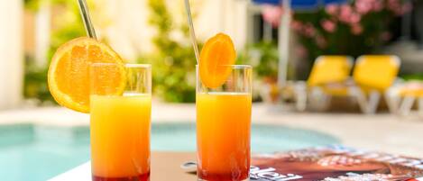 Two glasses of orange juice with orange slices on the rim, placed on a table near a poolside area with yellow chairs and a blue umbrella in the background.