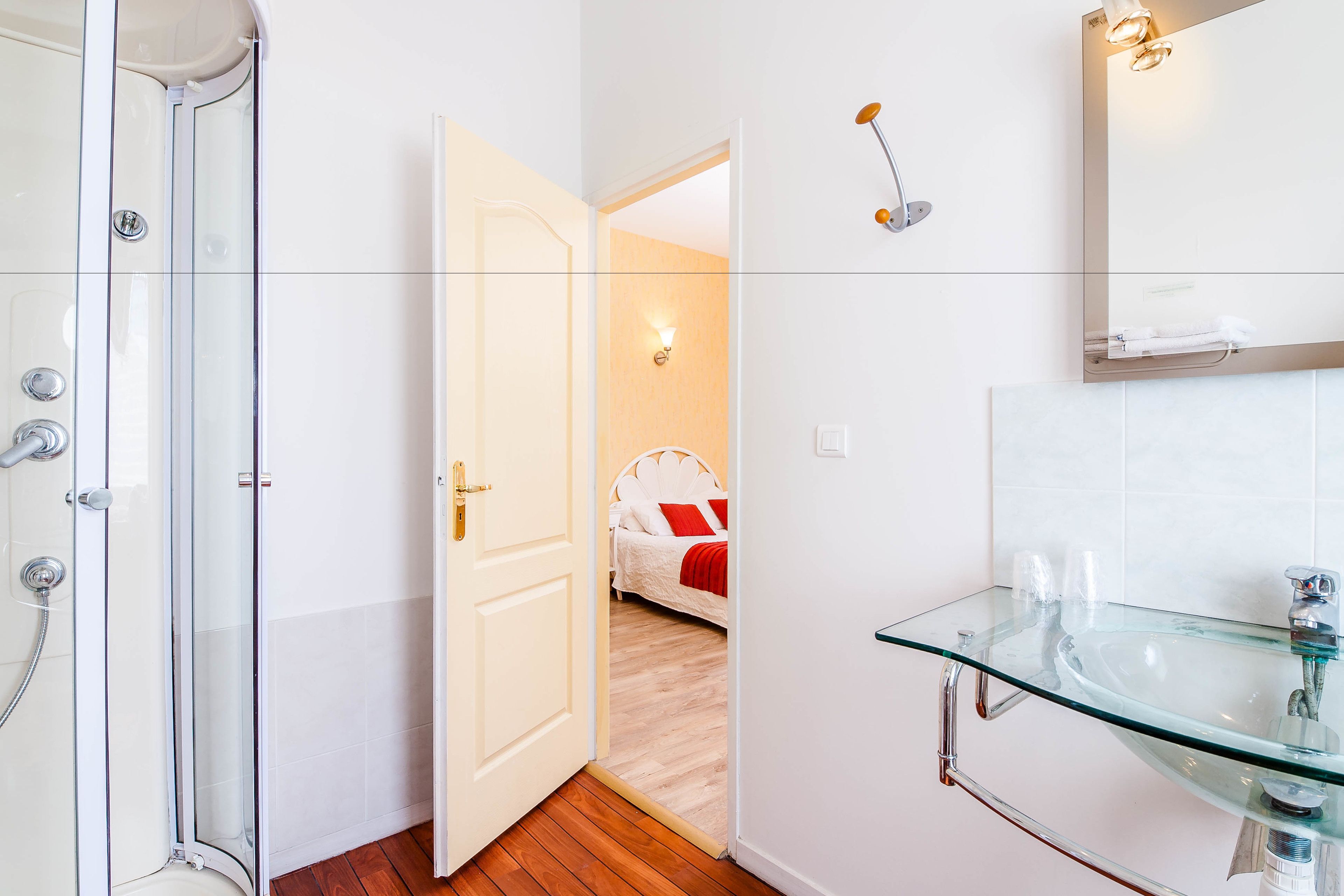 Ein modernes Badezimmer mit einer Duschkabine aus Glas, einem Waschbecken aus Glas und einem Bett, das durch eine offene Tür sichtbar ist.