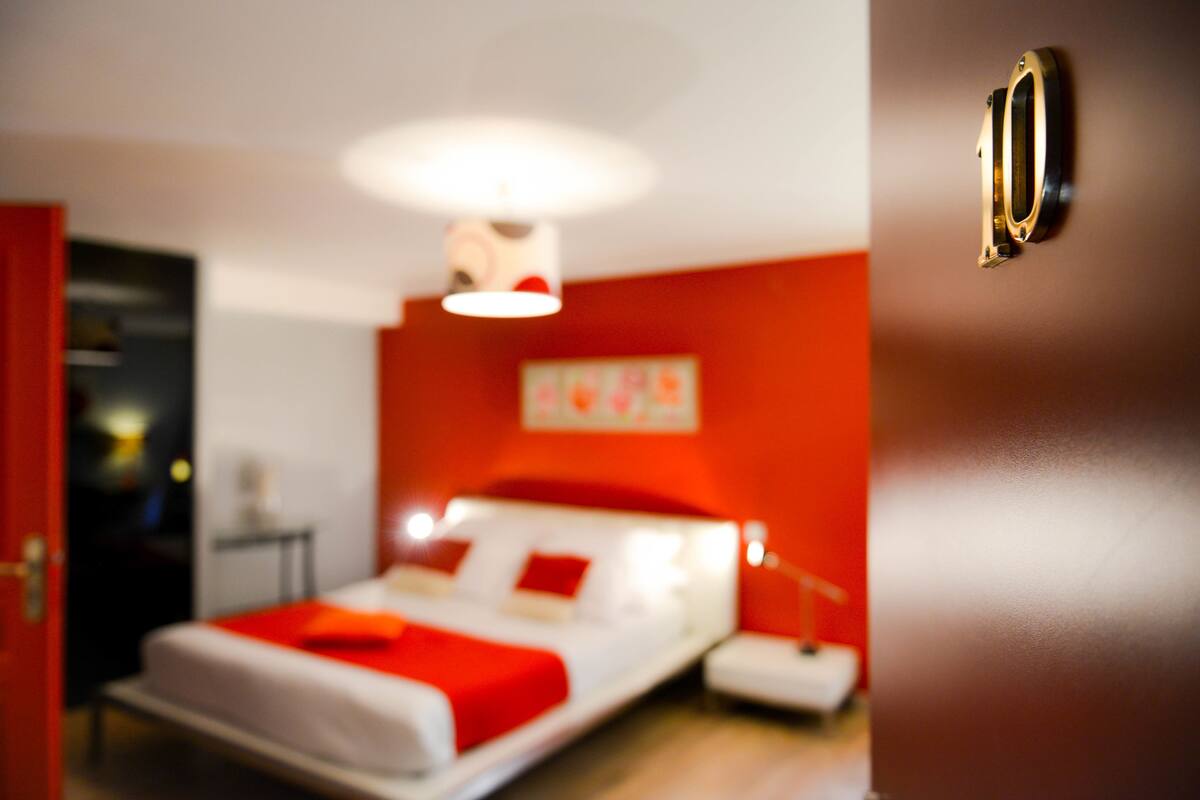 Un lit avec une couverture rouge, une table de chevet et un luminaire fixé au mur.