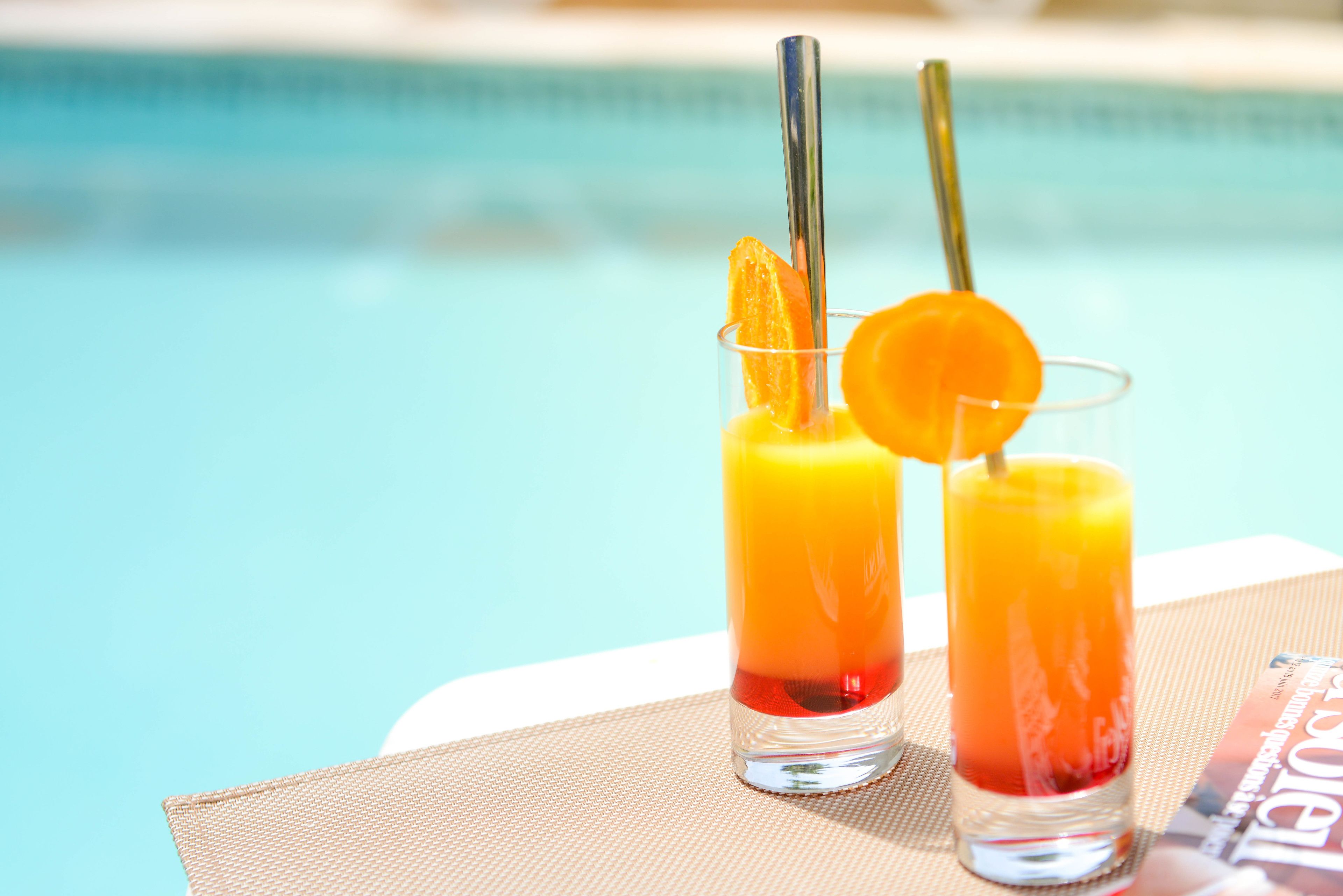 Dos vasos de jugo de naranja con pajitas y una rodaja de naranja sobre una mesa junto a una piscina.