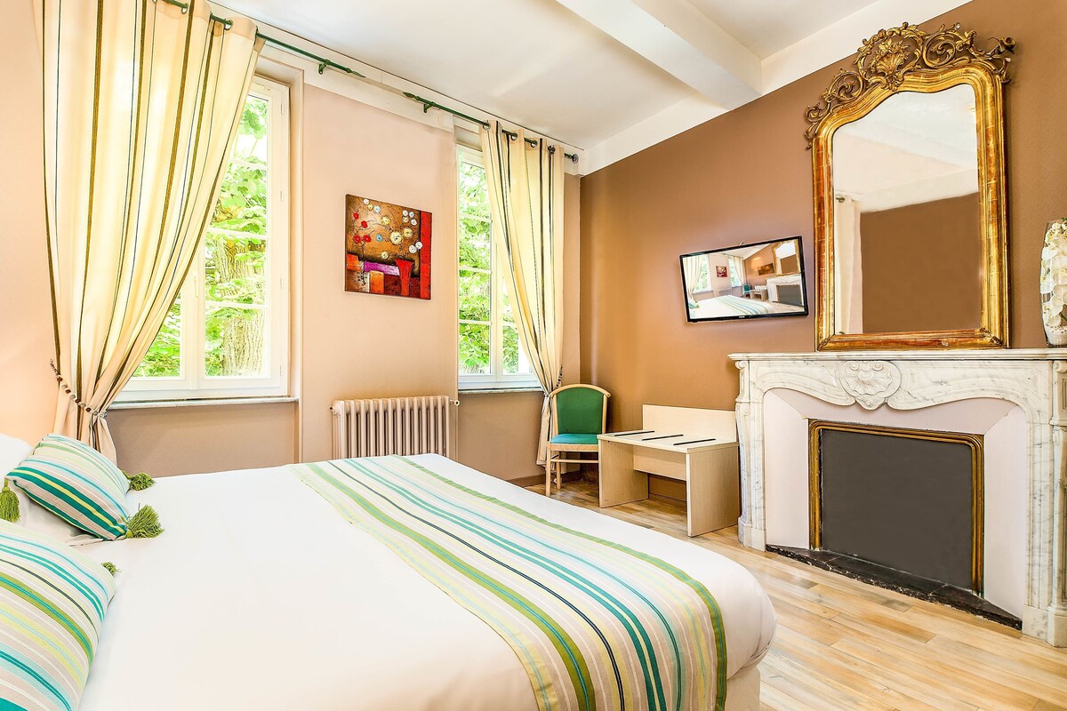 A hotel room with a bed, a desk, a chair, a fireplace, a large mirror, and a window with curtains.