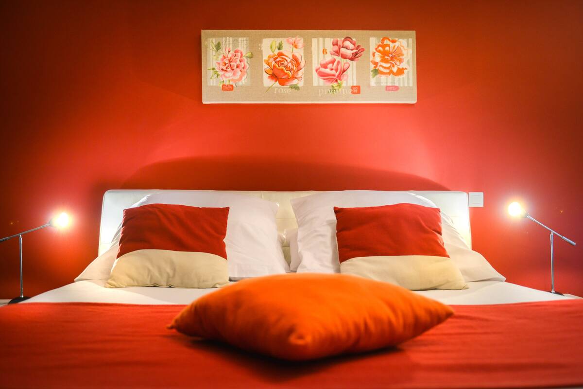 Un lit avec une literie rouge et blanche, deux oreillers rouges et un seul oreiller orange.