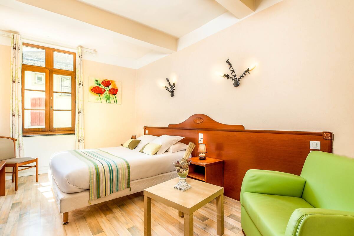 Une chambre d’hôtel avec un lit, une table de chevet, un fauteuil vert, une tête de lit en bois, une lampe fixée au mur et une fenêtre avec des rideaux.