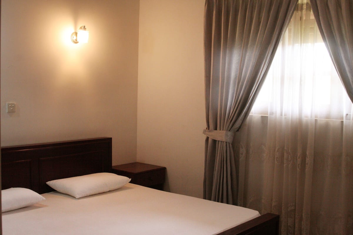 A hotel room with a bed, a nightstand, a wall-mounted light, and a window with curtains.