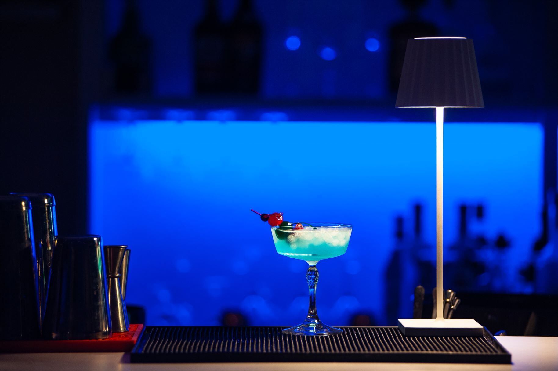 Un bar con una copa de cóctel sobre una bandeja, una lámpara y botellas al fondo.