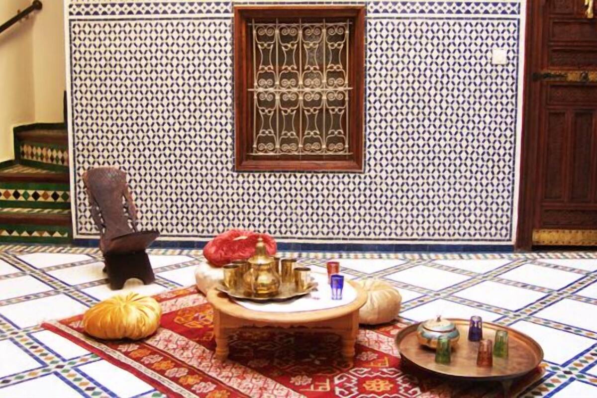 A traditional Moroccan room with patterned tiles, a wooden door, a small window with a metal grille, and a central rug with a table set with items.
