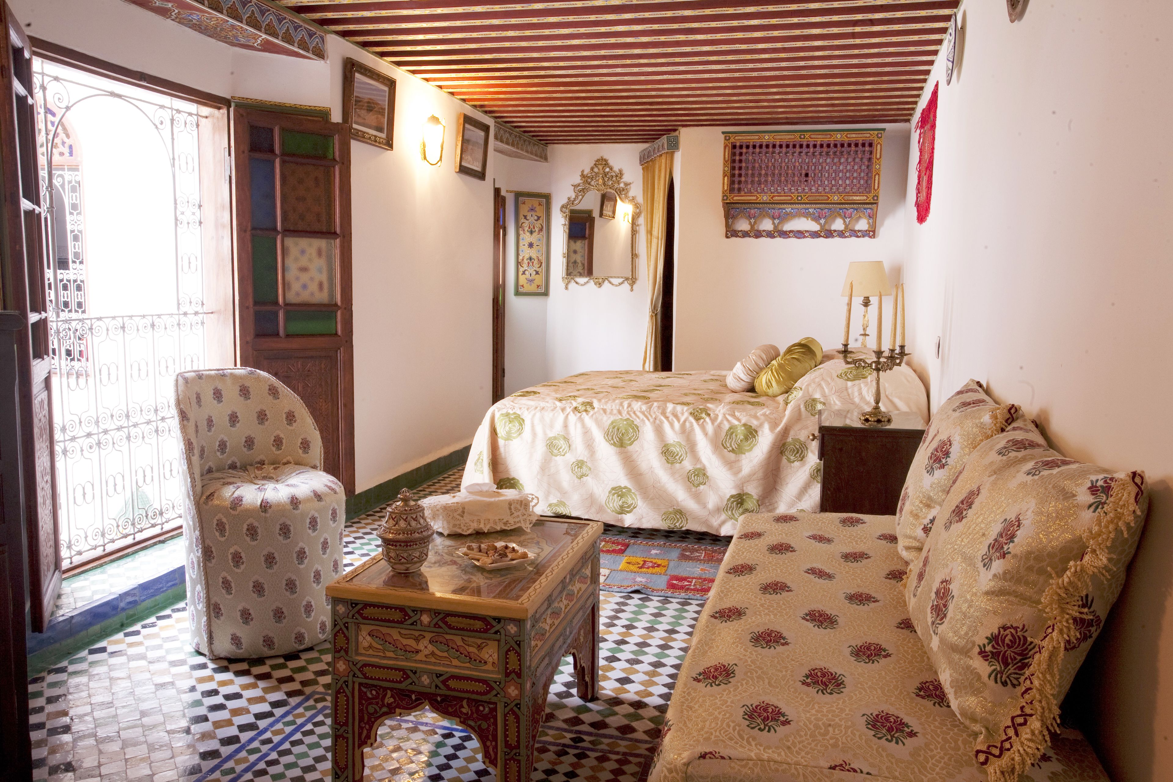 Una habitació marroquina tradicional amb un paviment motllejat, un llit amb colcha verd i blanca, un sofà amb dibuixos florals i una taula petita amb un objecte decoratiu.
