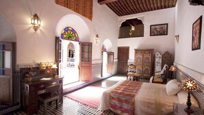 Quarto tradicional marroquino com piso quadriculado, cama com colcha estampada, escrivaninha com luminária e um pendurar decorativo na parede.