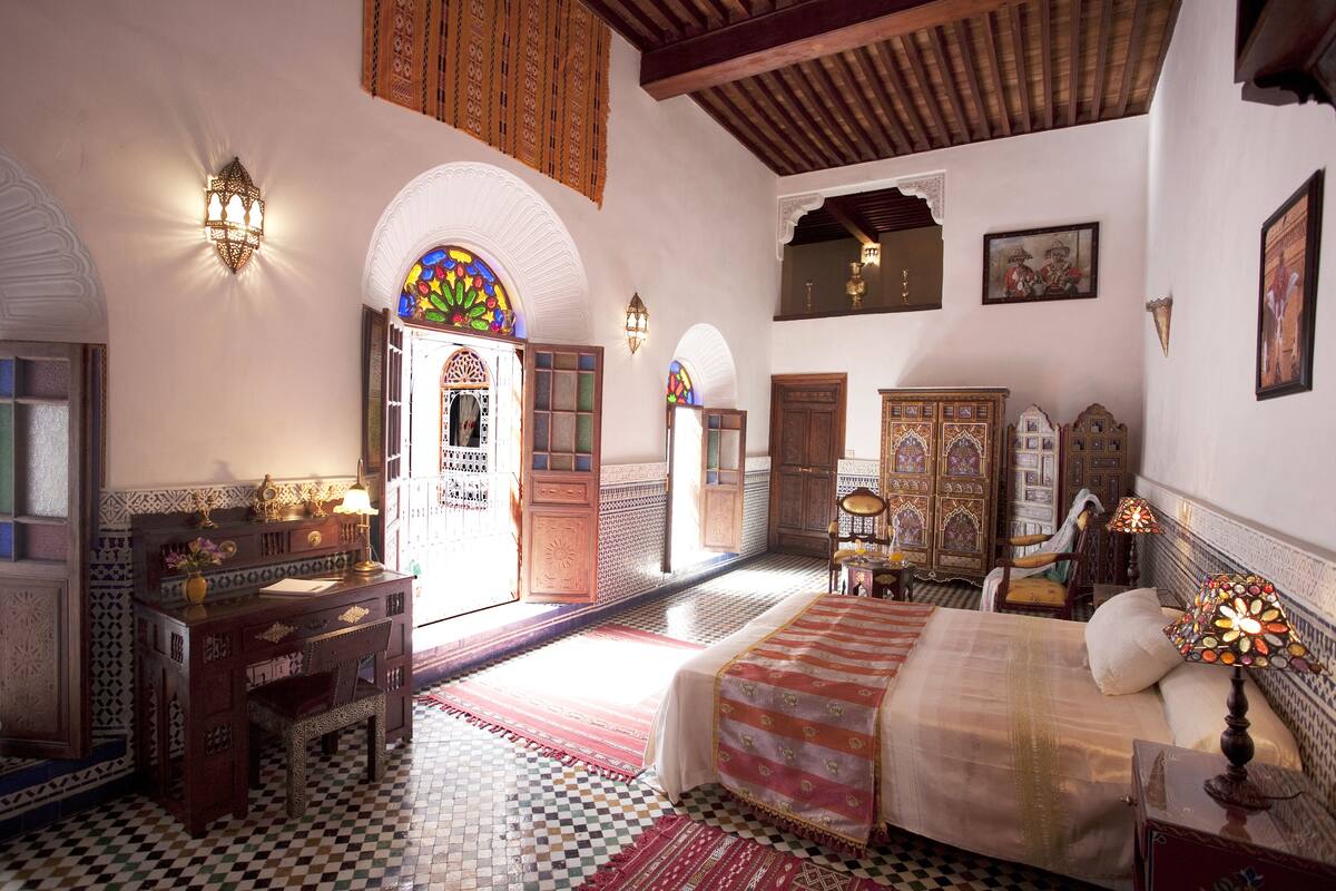 A traditional Moroccan room with a checkered floor, a bed with a patterned bedspread, a desk with a lamp, and a decorative wall hanging.